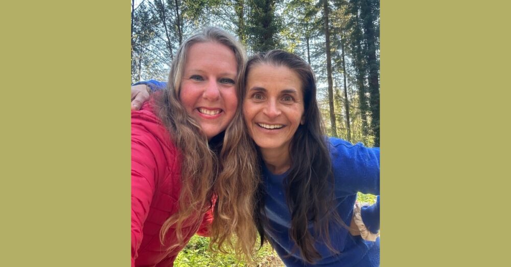 Two smiling women, one in a red jacket and the other in a blue sweater, taking a selfie in a sunlit forest.